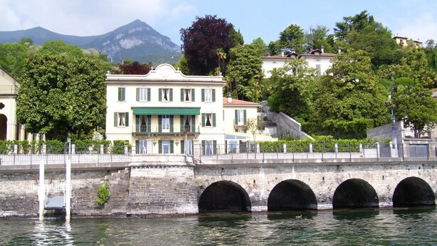 A stone bridge with three arches over a body of water, leading to a building with a green awning and multiple windows. - Villa La Mirabella (Griante)