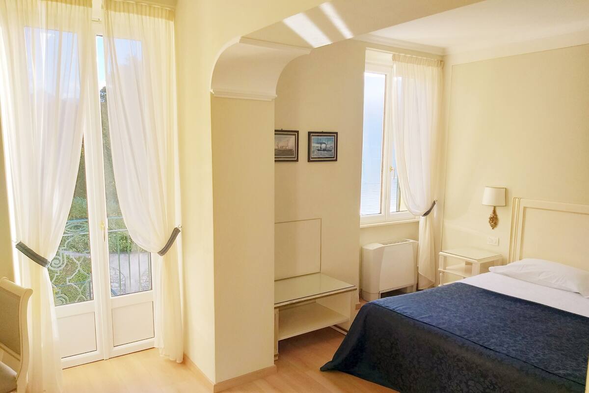 A hotel room with a bed, a desk, a chair, a window with curtains, and a view of greenery outside.