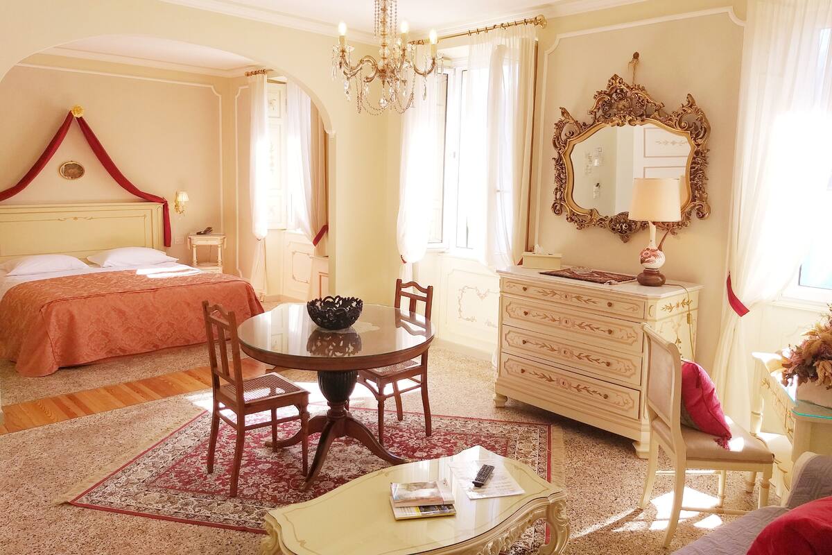 A hotel room with a bed, a dining table, a chandelier, a dresser, and a mirror.
