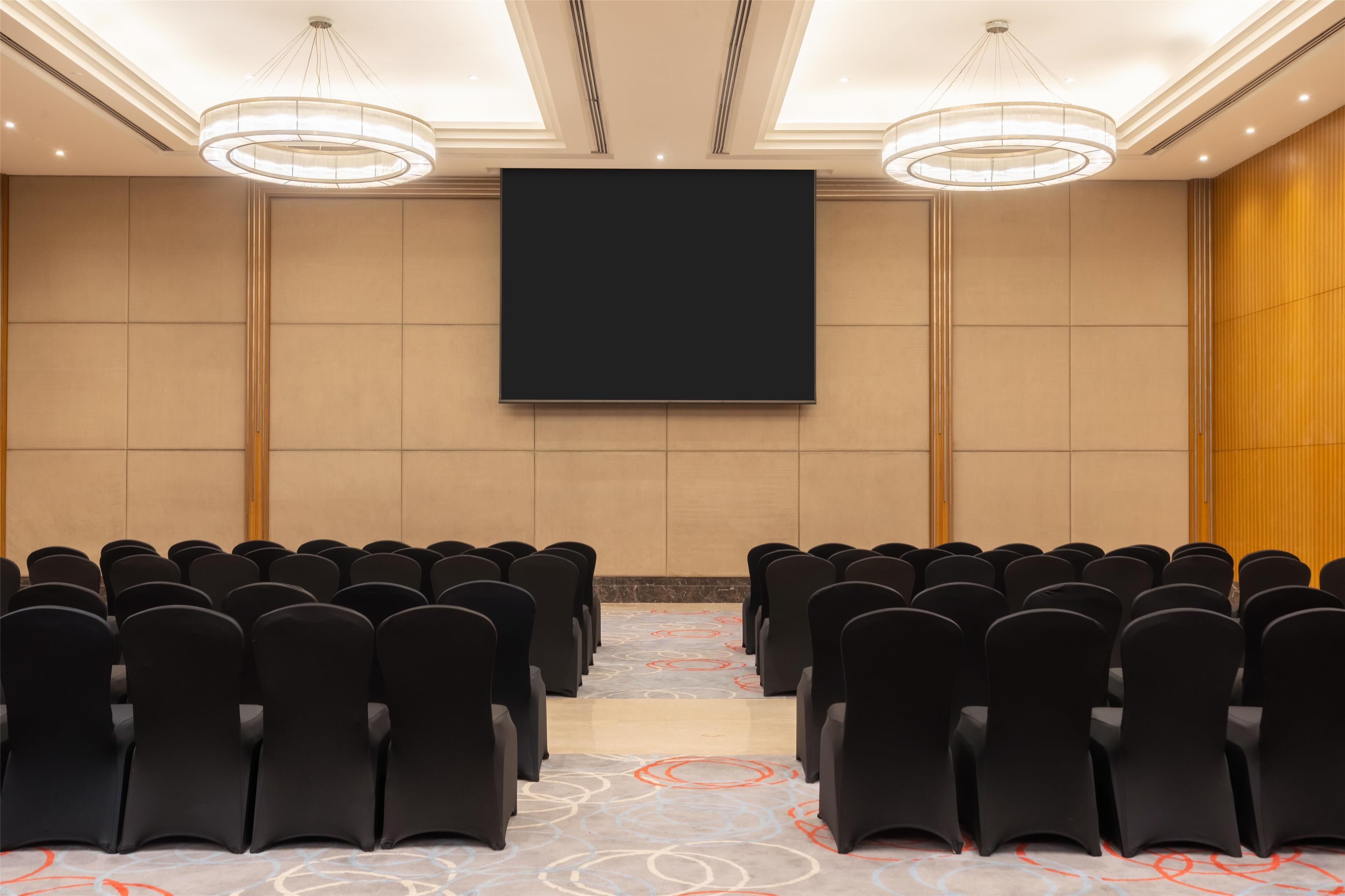 A conference room with rows of black chairs, a large screen, and a patterned carpet.