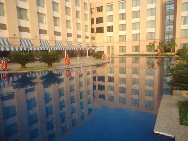 A hotel pool with clear water, surrounded by lounge chairs and umbrellas, reflecting the building's facade.