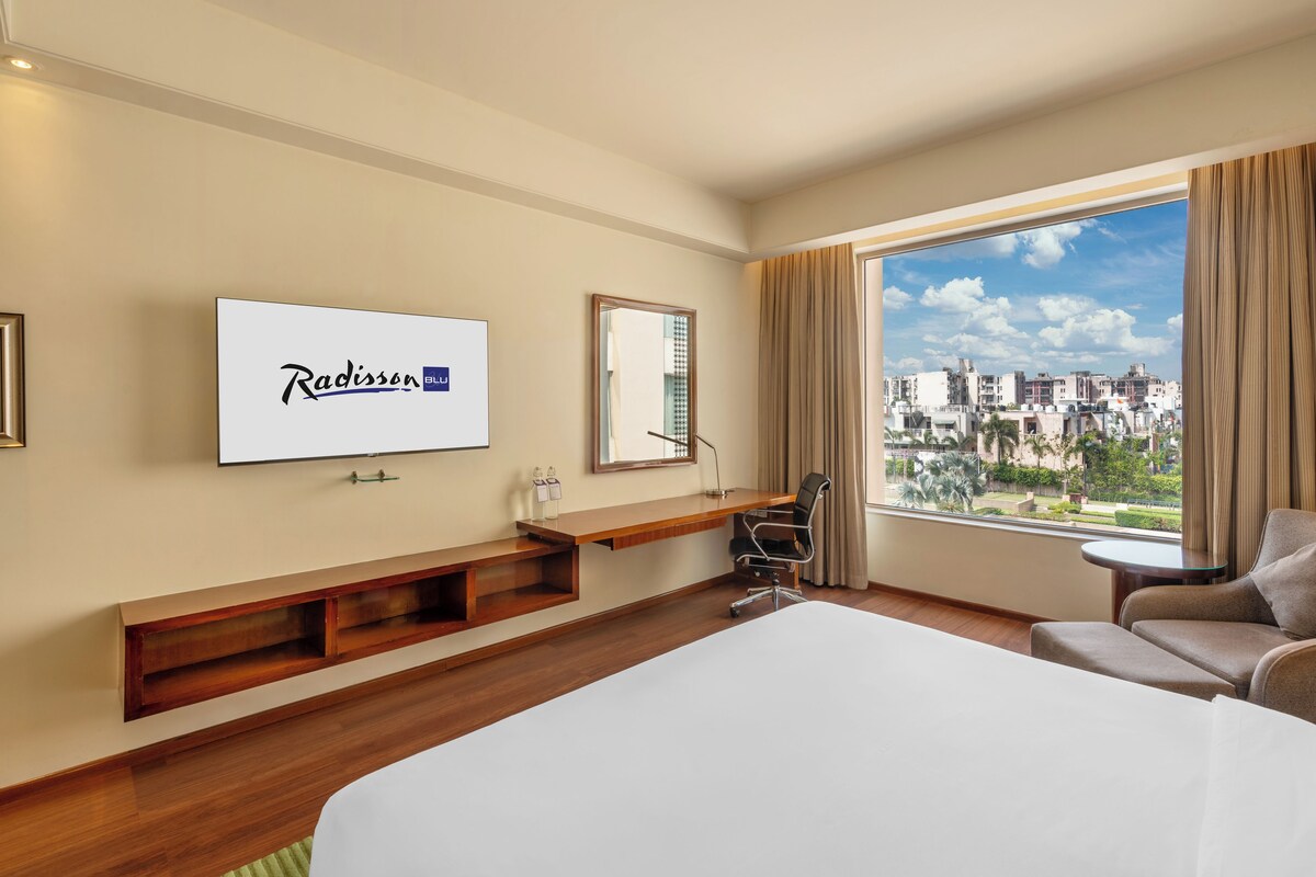A hotel room with a bed, a desk, a chair, a TV mounted on the wall, and a large window offering a city view.
