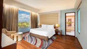 A hotel room with a large bed, a desk, a chair, a small table, and a view of greenery outside.