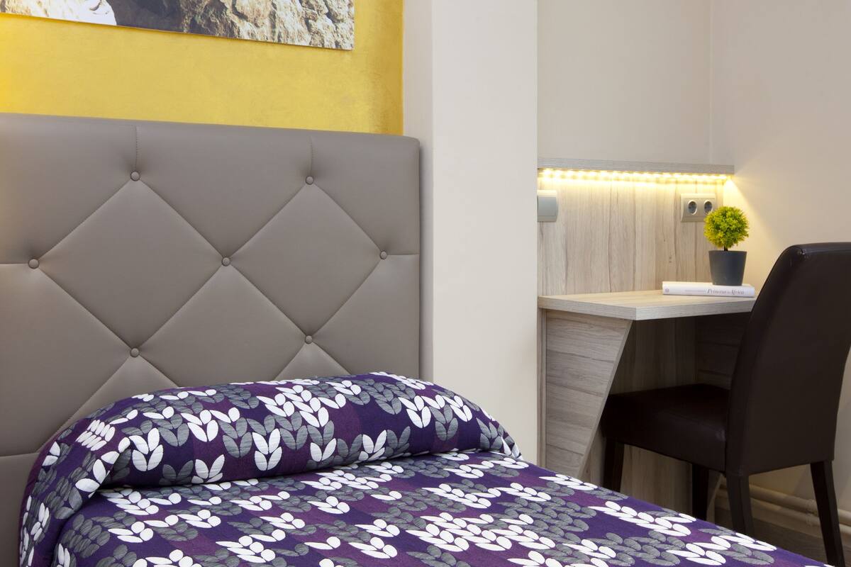 A hotel room with a tufted headboard, a bed with a purple floral bedspread, a desk with a chair, and a small potted plant.