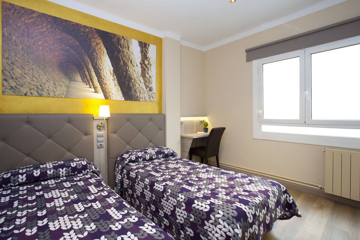 A hotel room with two beds, a desk, a chair, a large window, and a painting on the wall.