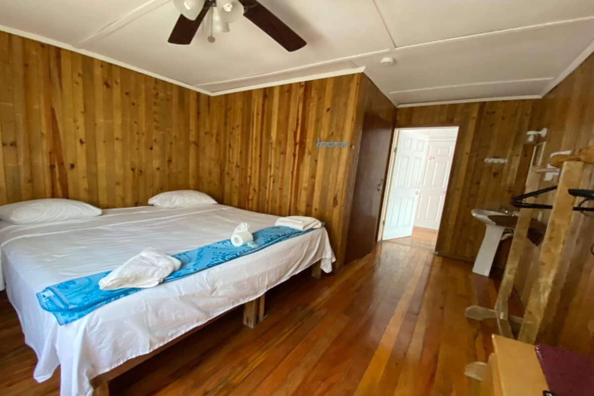 A wooden-paneled room with a bed, a ceiling fan, and a bathroom with a sink and mirror.