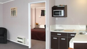 Ein kompaktes Hotelzimmer mit Küchenzeile, einem Bett mit roter Bettdecke, einem kleinen Esstisch und einem Stuhl.