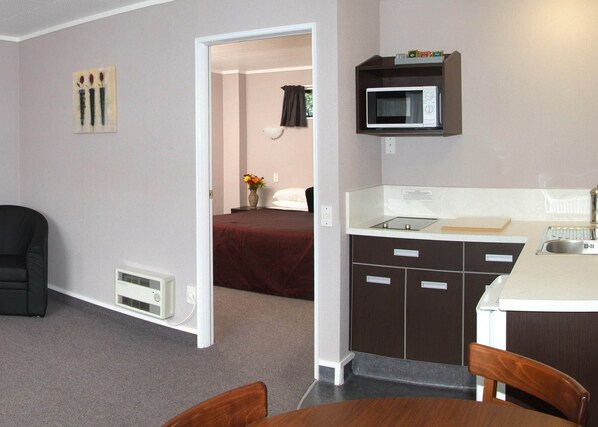 A compact hotel room with a kitchenette, a bed with a red cover, a small dining table, and a chair. - Rose City Motel (Palmerston North)