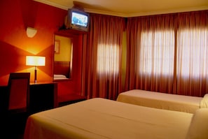 A hotel room with two beds, a television on a stand, a desk with a lamp, and red curtains.