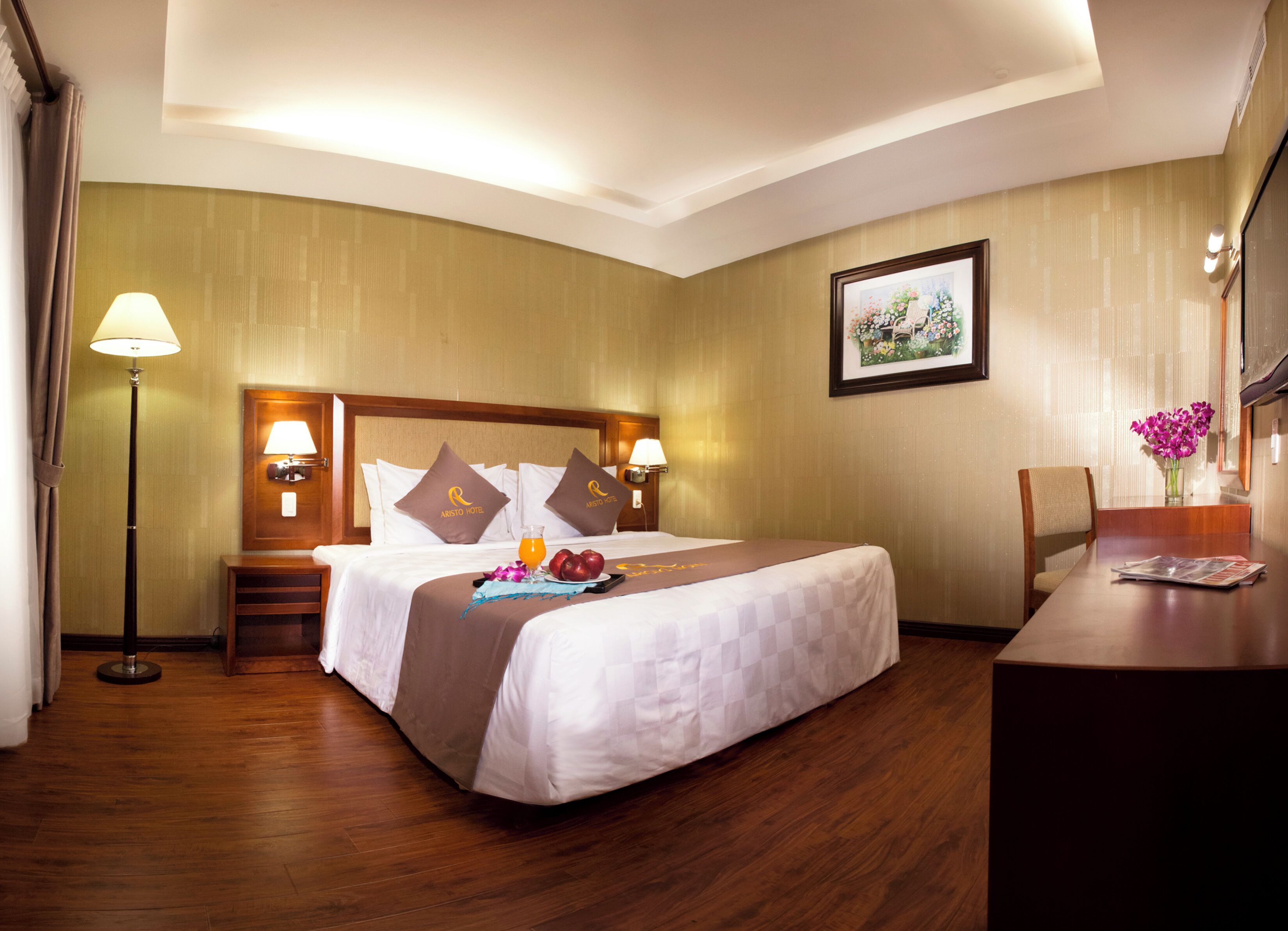 Quarto de hotel com uma cama grande, uma escrivaninha, uma cadeira, um abajur e um quadro na parede.
