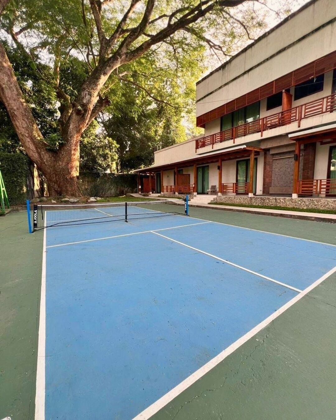 An outdoor tennis court with a net, surrounded by trees and a building with balconies.