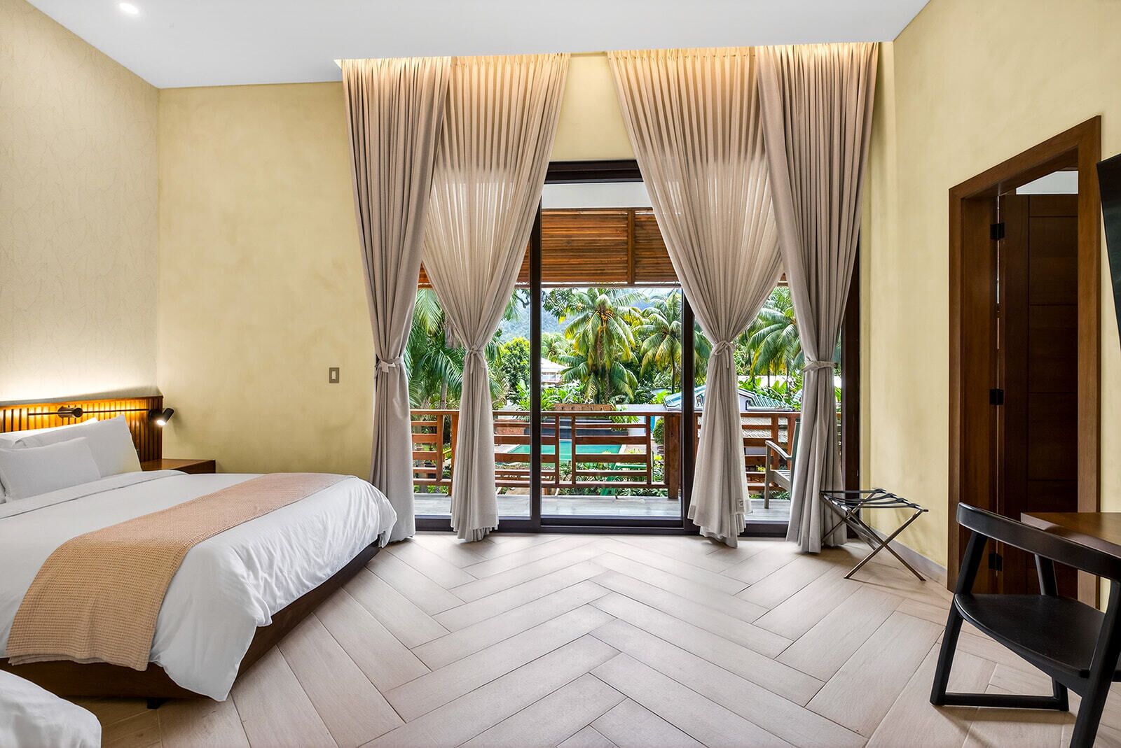 Une chambre d’hôtel avec un lit, un bureau, une chaise et une vue sur les palmiers par la fenêtre.