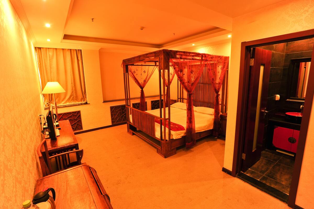 A four-poster bed with a canopy, a desk with a lamp, and a bathroom with a red sink.
