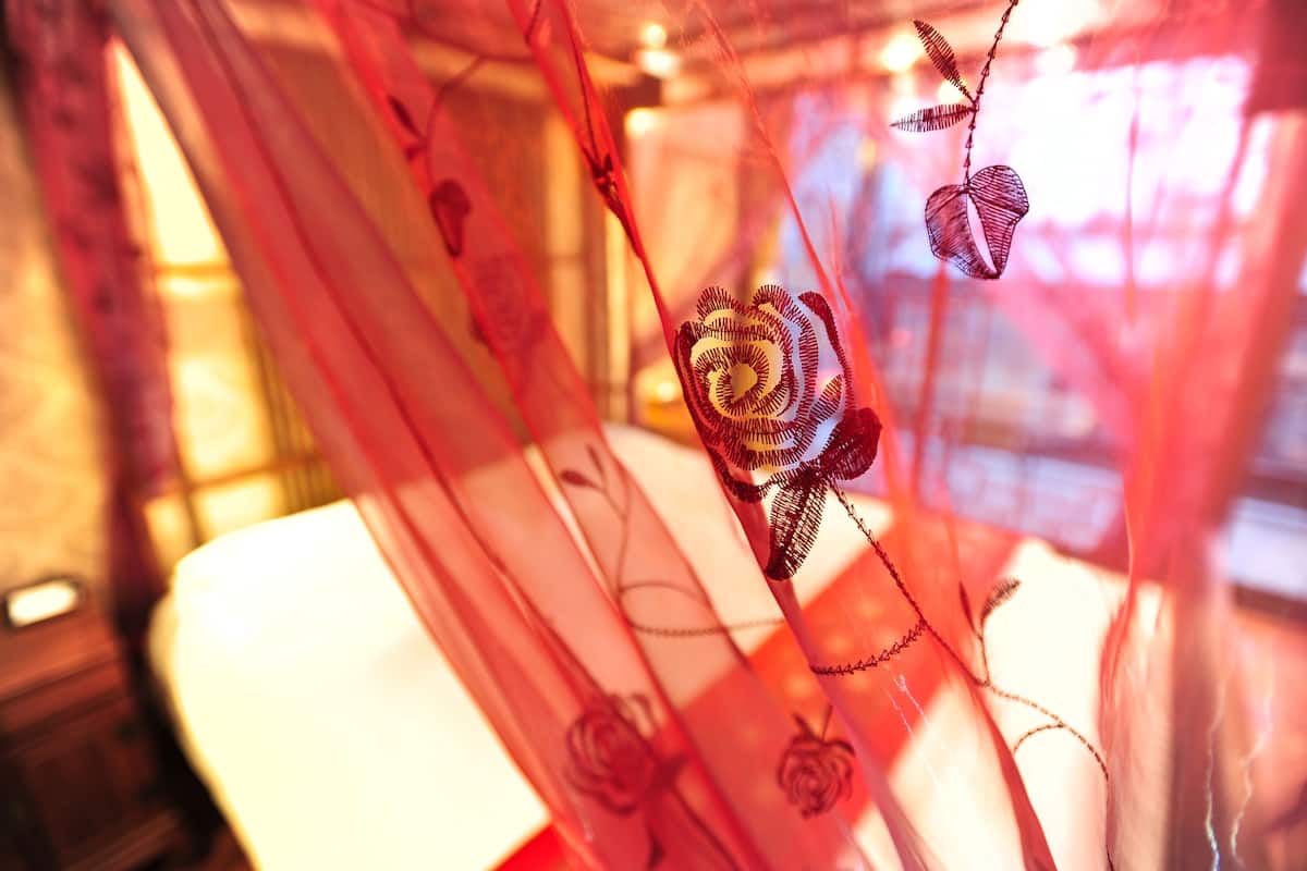 A room with a bed covered by a sheer red curtain with floral embroidery.