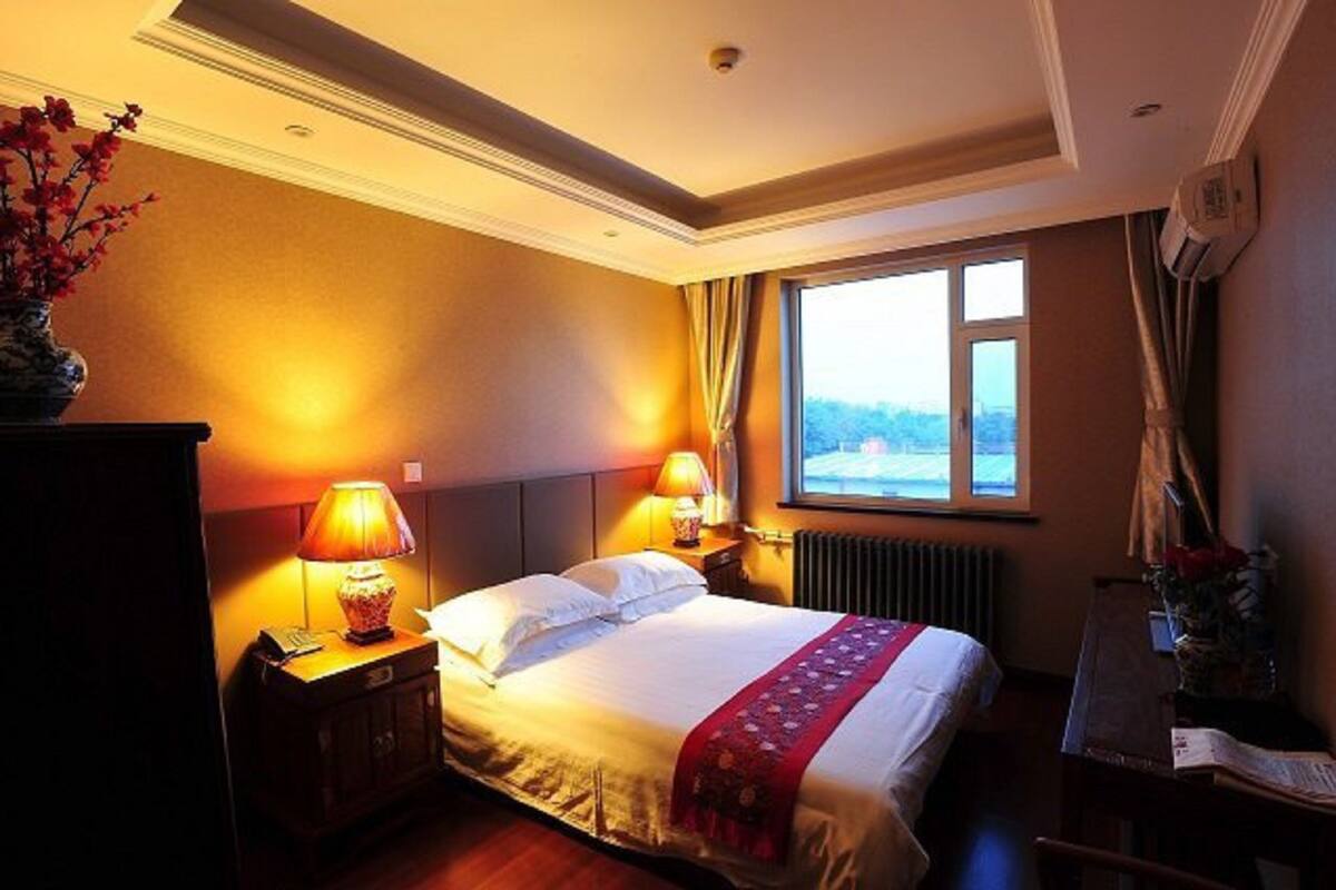 A hotel room with a large bed, two bedside lamps, a radiator, a window with a view, and a desk with a lamp.