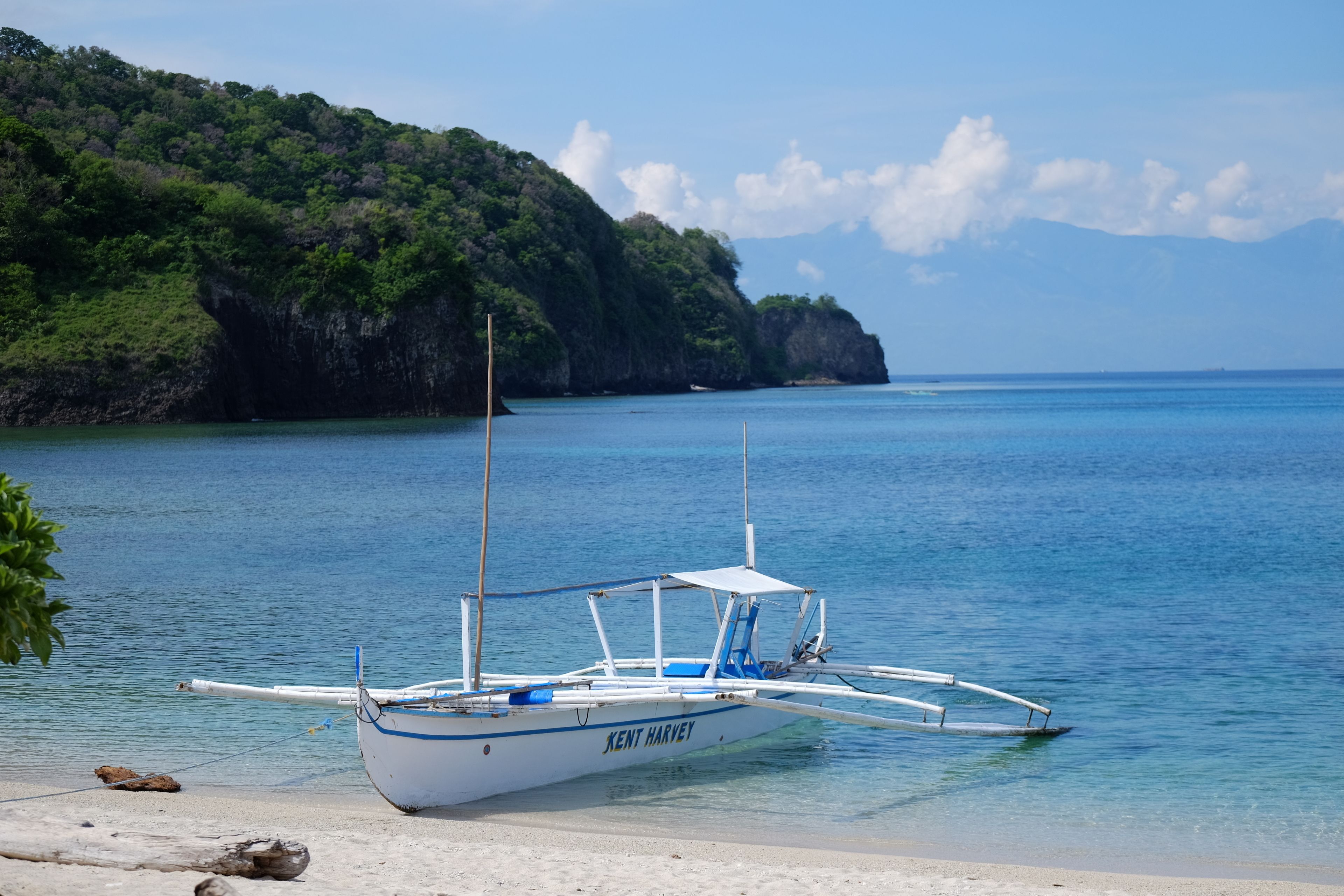 A boat named "Kent Harvey" anchored near a beach with clear blue water and a forested hill in the background.