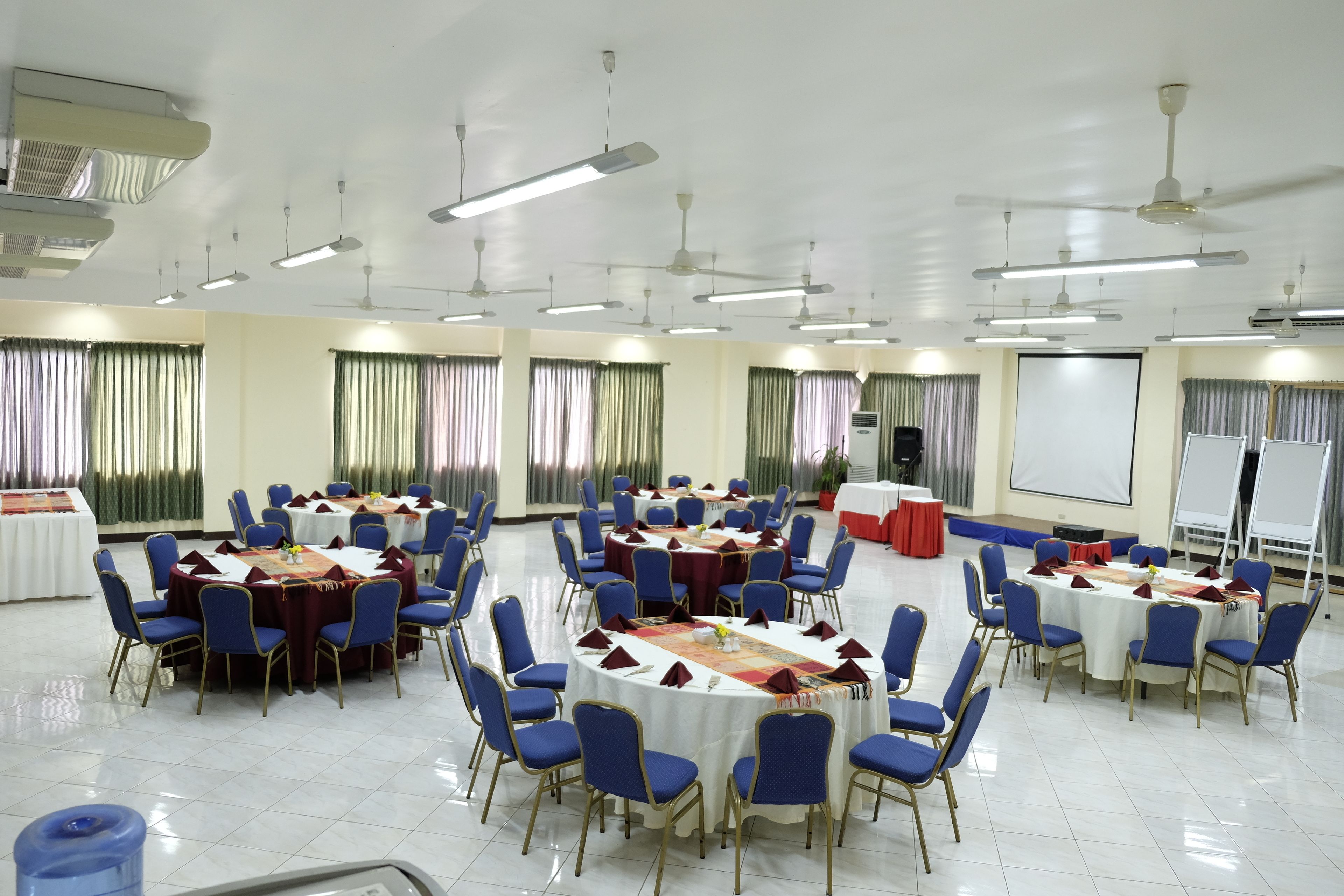 A conference room with multiple tables and chairs, ceiling fans, and a projector screen.