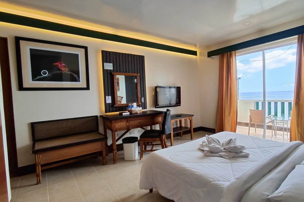 A hotel room with a bed, a desk, a chair, a TV, and a balcony with a view of the ocean.