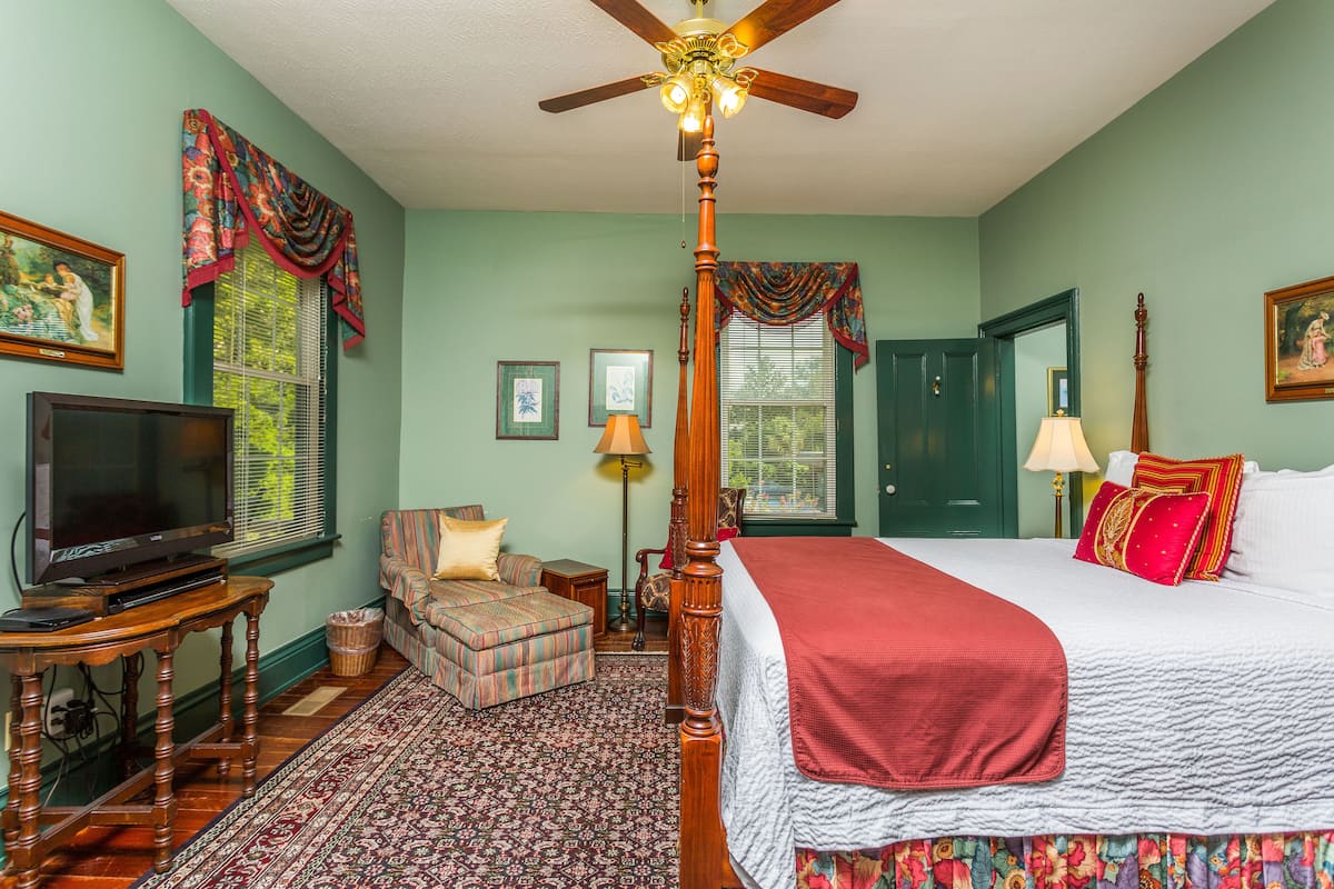 A bedroom with a four-poster bed, a television, a chair, and a large window with curtains.