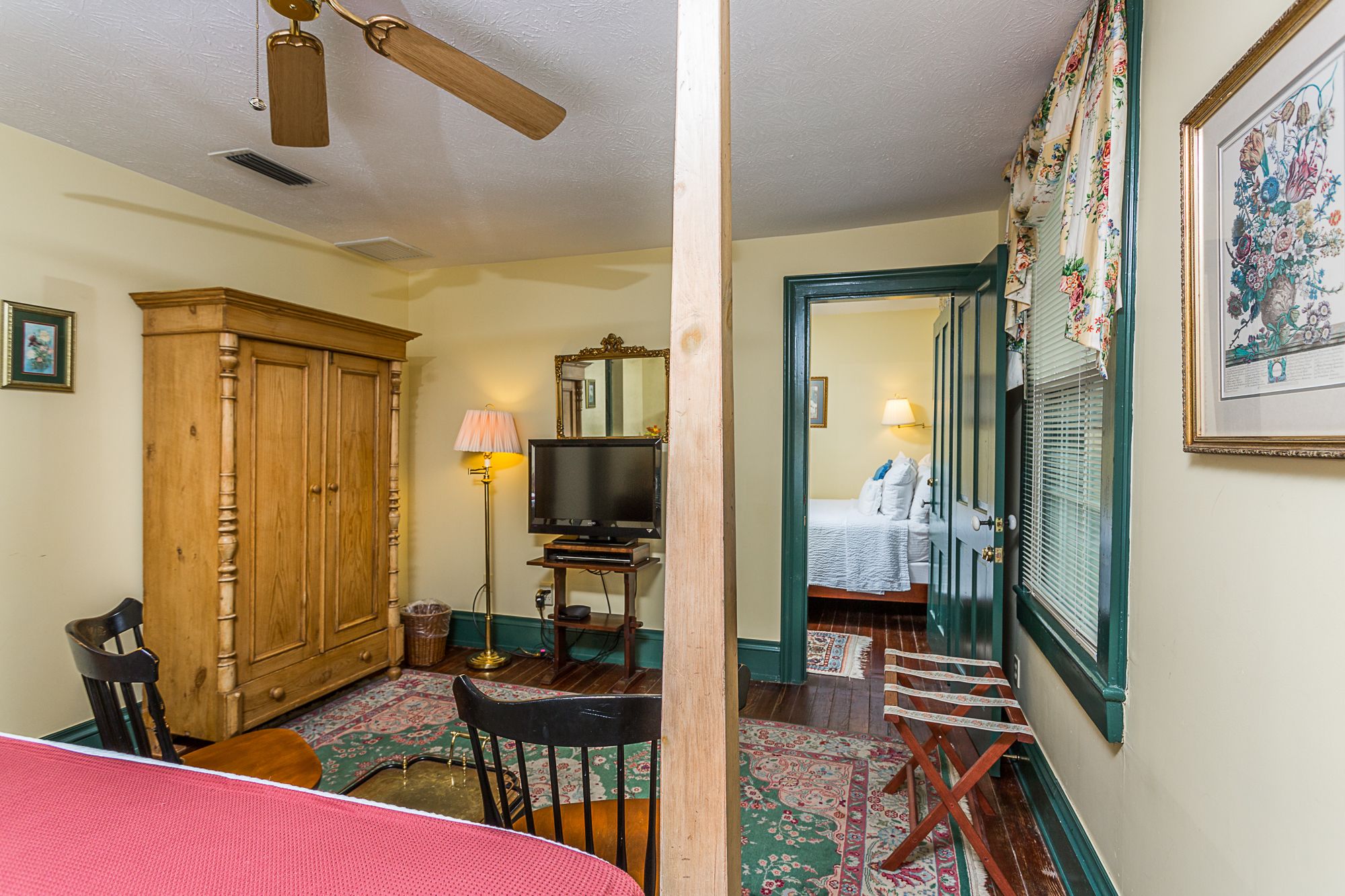 A hotel room with a wooden wardrobe, a television, a lamp, a bed with white linens, and a floral painting on the wall.