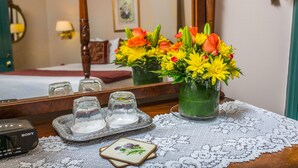 Quarto de hotel com uma cama, uma mesa de cabeceira com um vaso de flores, um apoio para bebidas com copos e um despertador Sony.