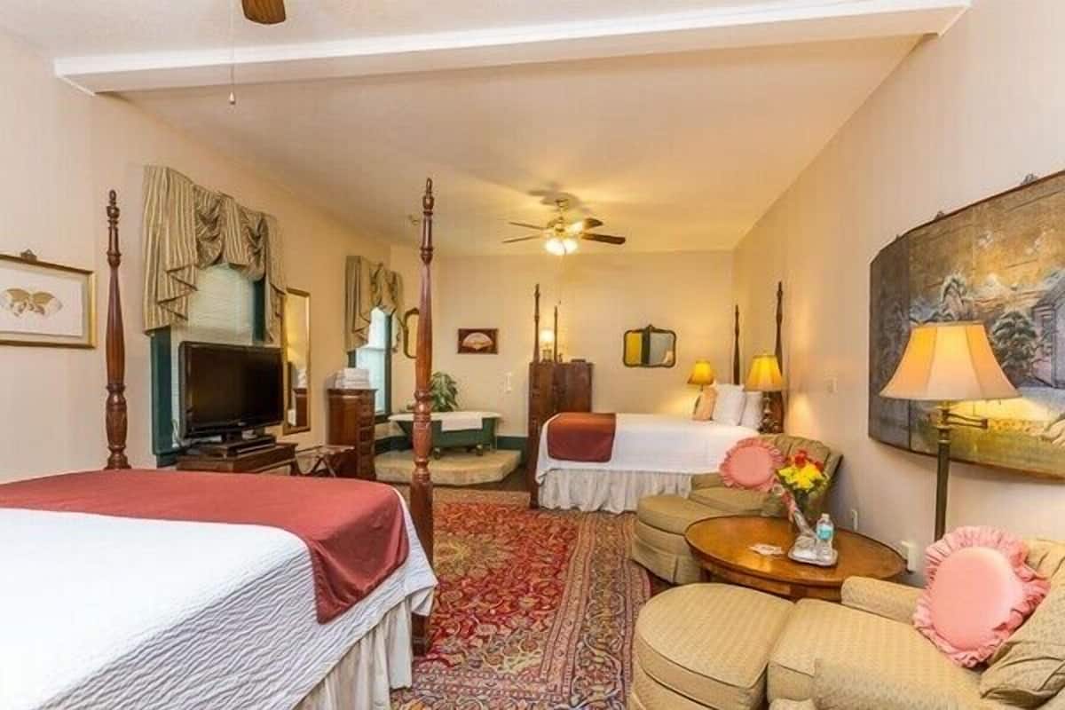 A hotel room with a large bed, a smaller bed, a sitting area with a sofa and armchairs, a television, a ceiling fan, and a painting on the wall.