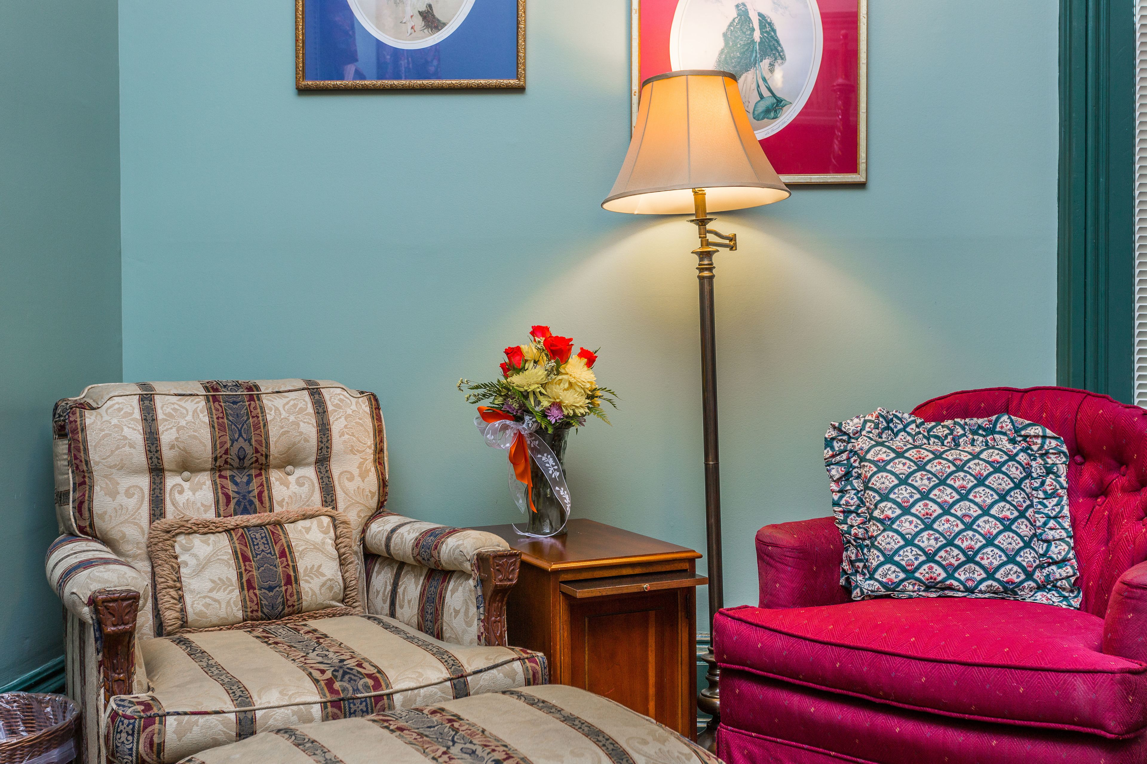 Een kamer met een gestreept fauteuil, een rode bank met een gekleurd kussen, een vloerlamp en een vaas met bloemen.