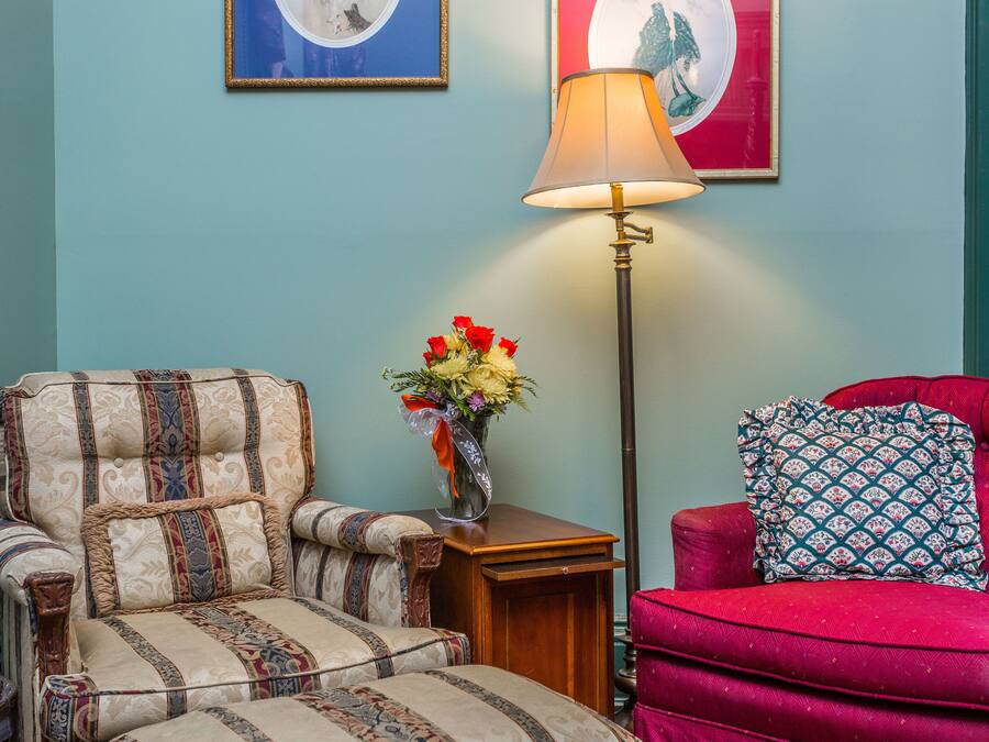 A room with a patterned armchair, a red sofa with a patterned pillow, a floor lamp, and a vase of flowers.