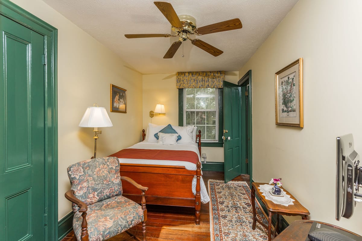 A bedroom with a wooden bed, a chair, a table with a vase, and a ceiling fan.