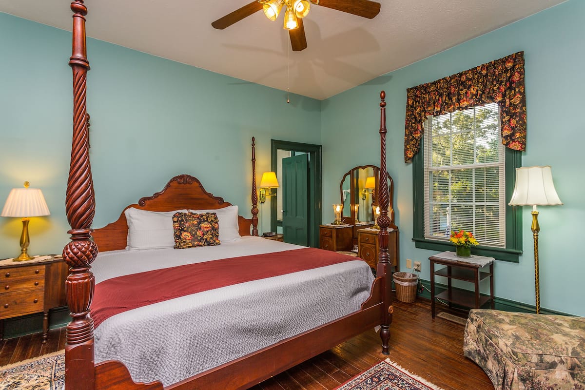 A four-poster bed with a wooden headboard, a wooden nightstand with a lamp, a dresser with a mirror, a window with a floral valance, and a small table with a lamp.