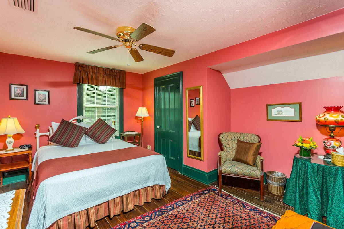 A bedroom with a bed, a chair, a table with a lamp, a mirror, and a ceiling fan.