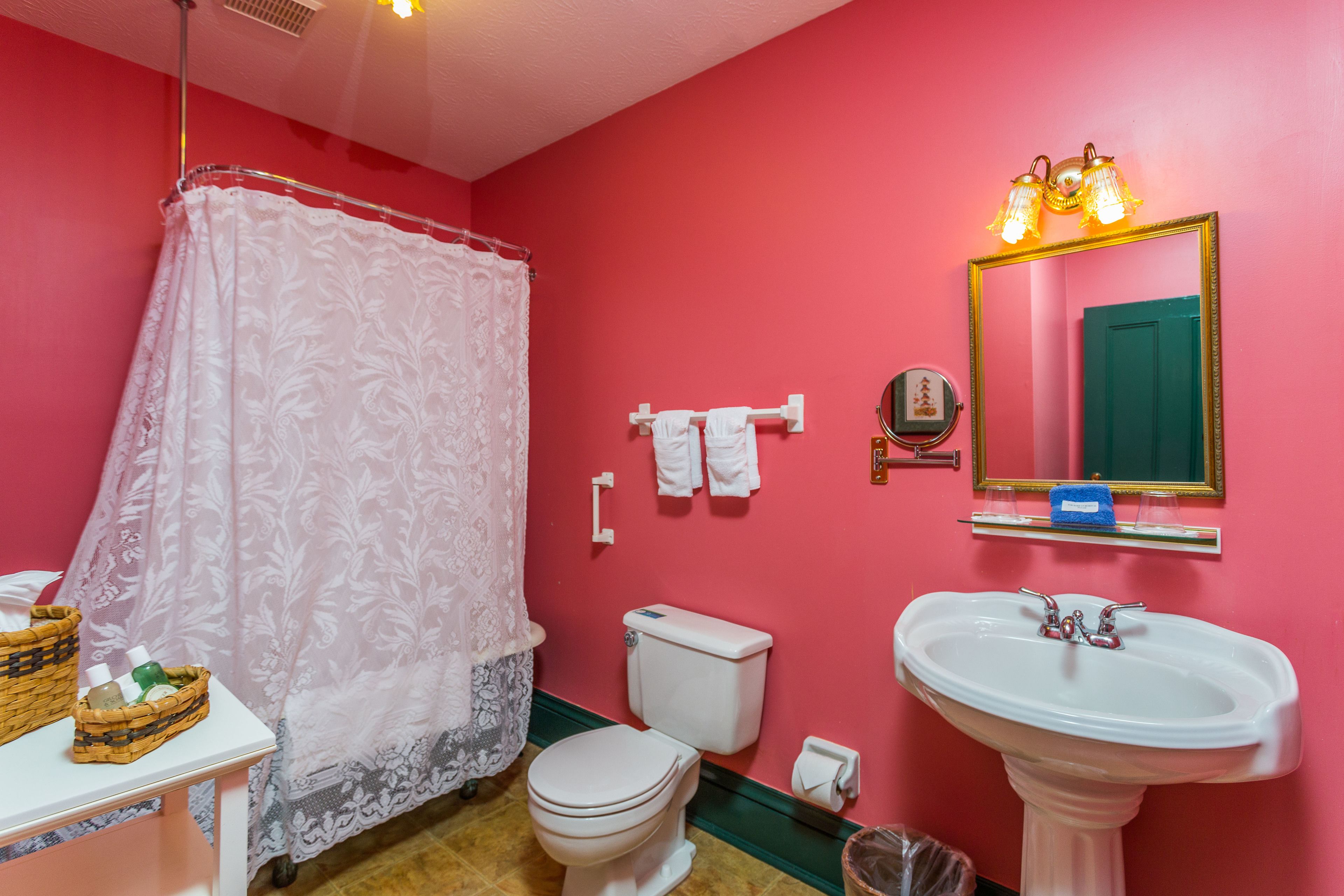 A bathroom with a pink wall, white toilet, white sink, and a shower with a lace curtain.