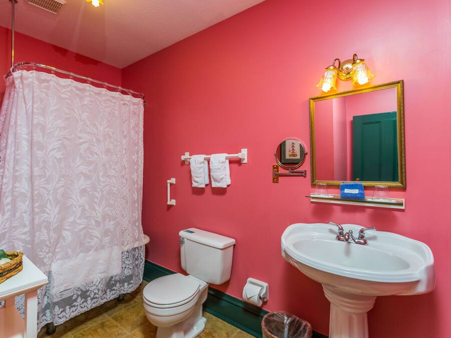 Casa de banho com parede cor-de-rosa, sanita branca, lavatório branco e chuveiro com cortina de renda.