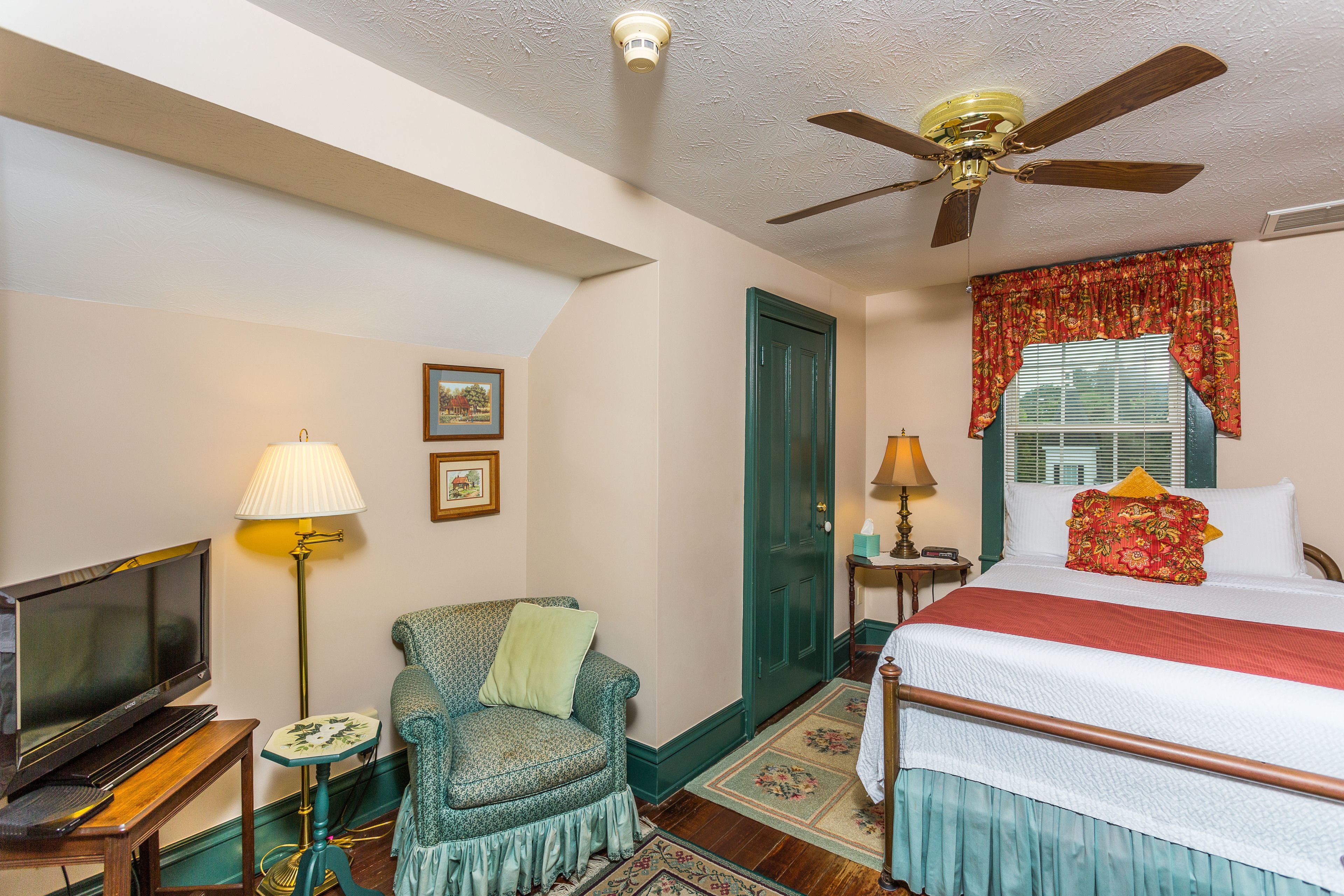 A bedroom with a bed, a television, a chair, a lamp, and a ceiling fan.