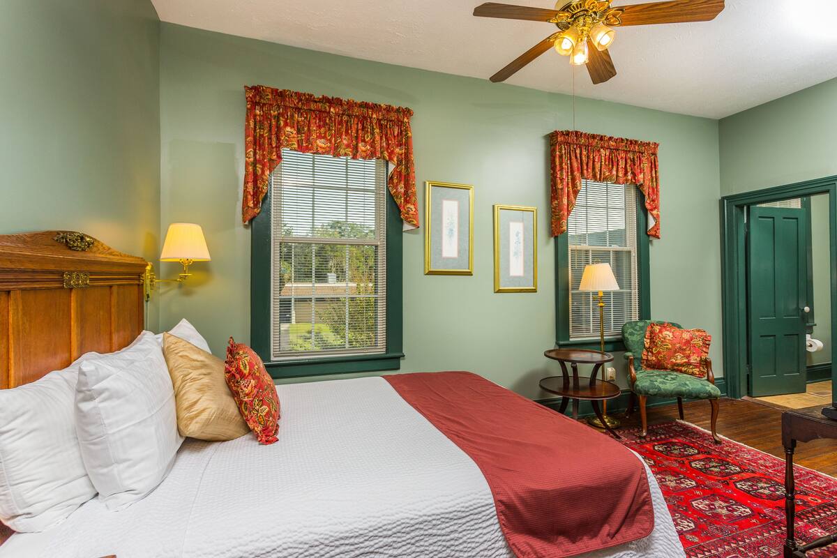 A bedroom with a bed, two windows with curtains, a chair, a table, and a door.