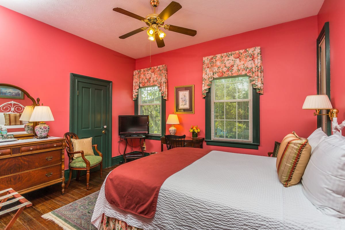 A bedroom with a bed, dresser, chair, television, and windows with floral curtains.