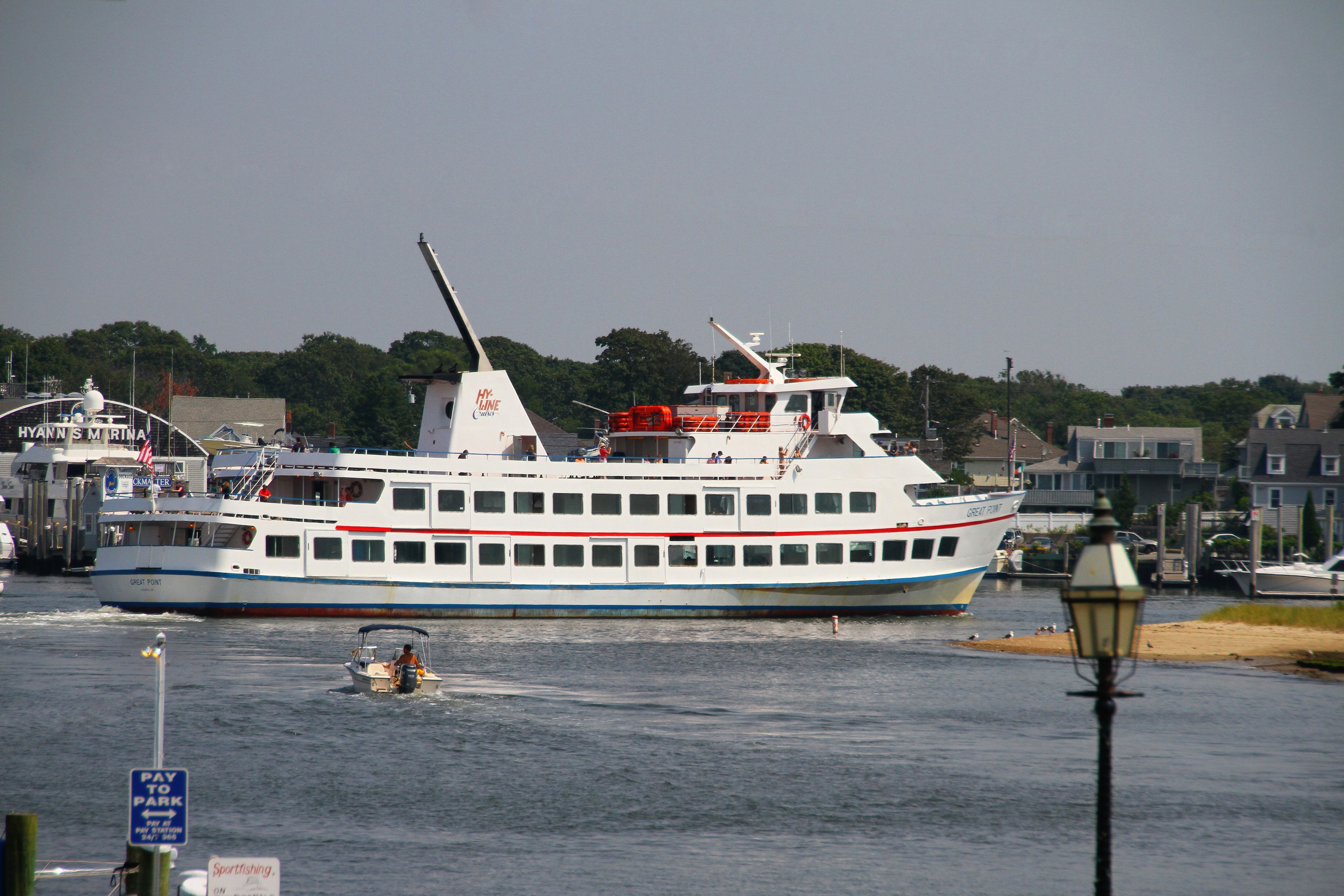 A large ferry with multiple decks and a flag, docked at a port with buildings and a lamp post in the foreground.