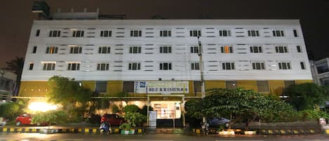 A hotel with a sign reading "Quality Inn" and "Bez Krishna." It has multiple floors, a well-lit entrance, and is surrounded by greenery.