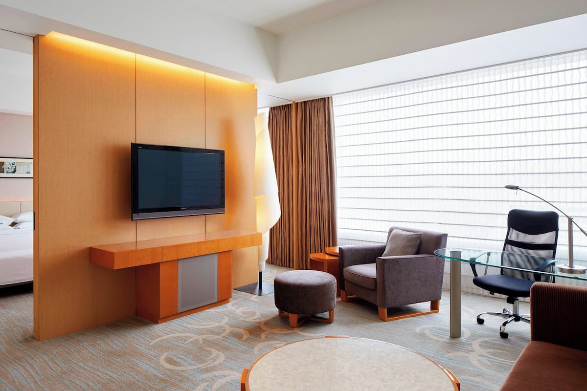 A modern hotel room with a flat-screen TV mounted on a wooden panel, a sofa, a chair, and a glass coffee table.