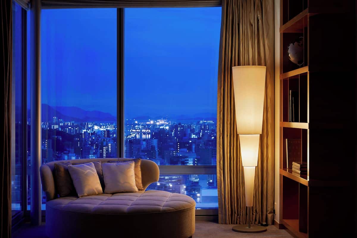 A modern living room with a large window, a sofa, a floor lamp, and a bookshelf.