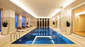 Una piscina interior moderna con un elemento acuático de agua azul cristalina, sillas de descanso y plantas en macetas.