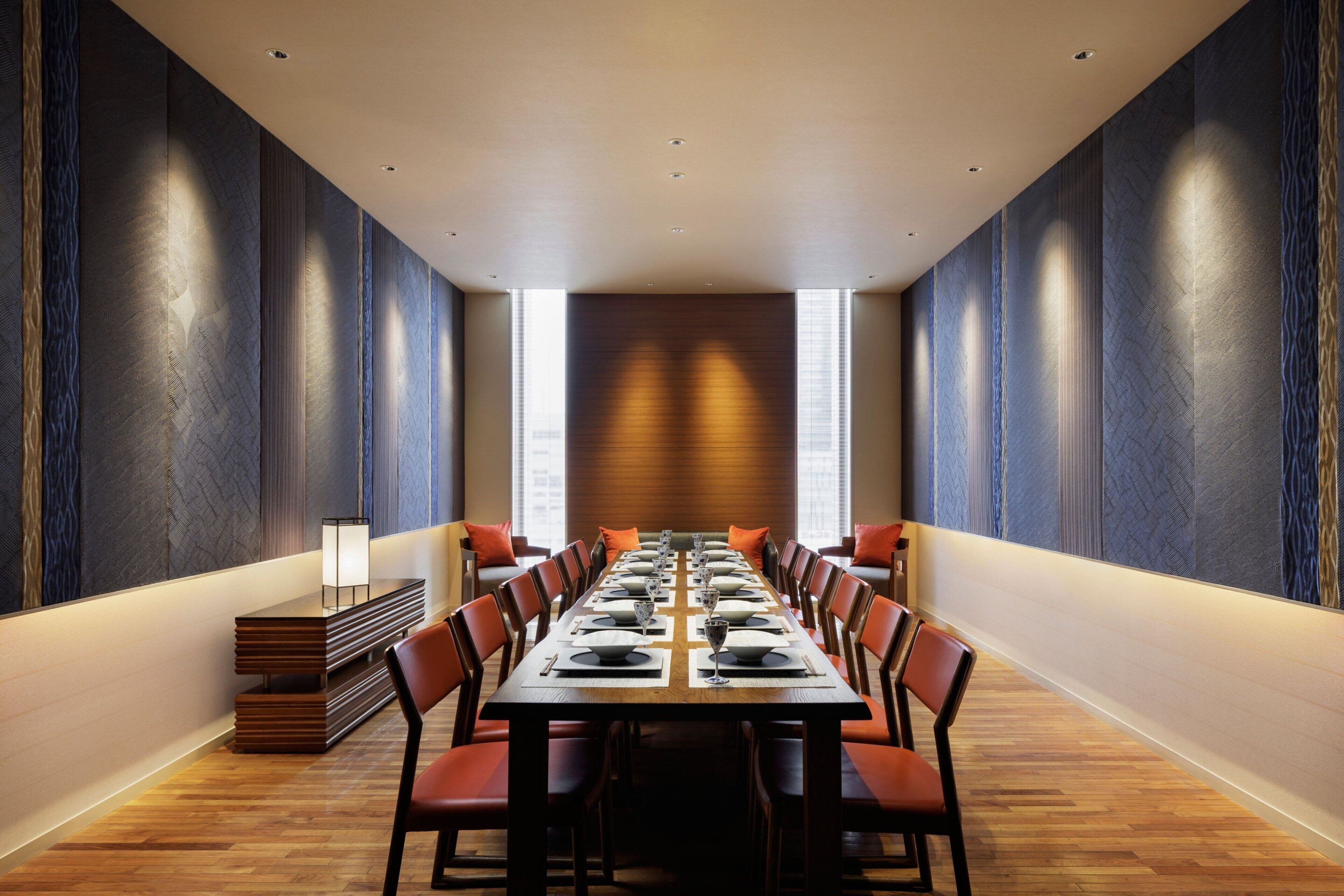 Un salón de conferencias con una larga mesa dispuesta para una reunión, rodeado de sillas, con un diseño moderno y ventanas amplias.