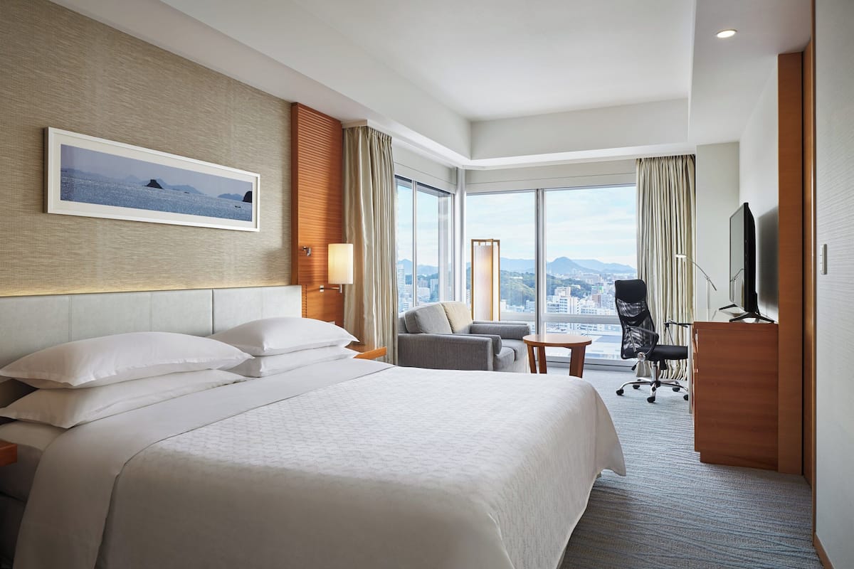A hotel room with a large bed, a desk, a chair, a TV, and a view of the city.