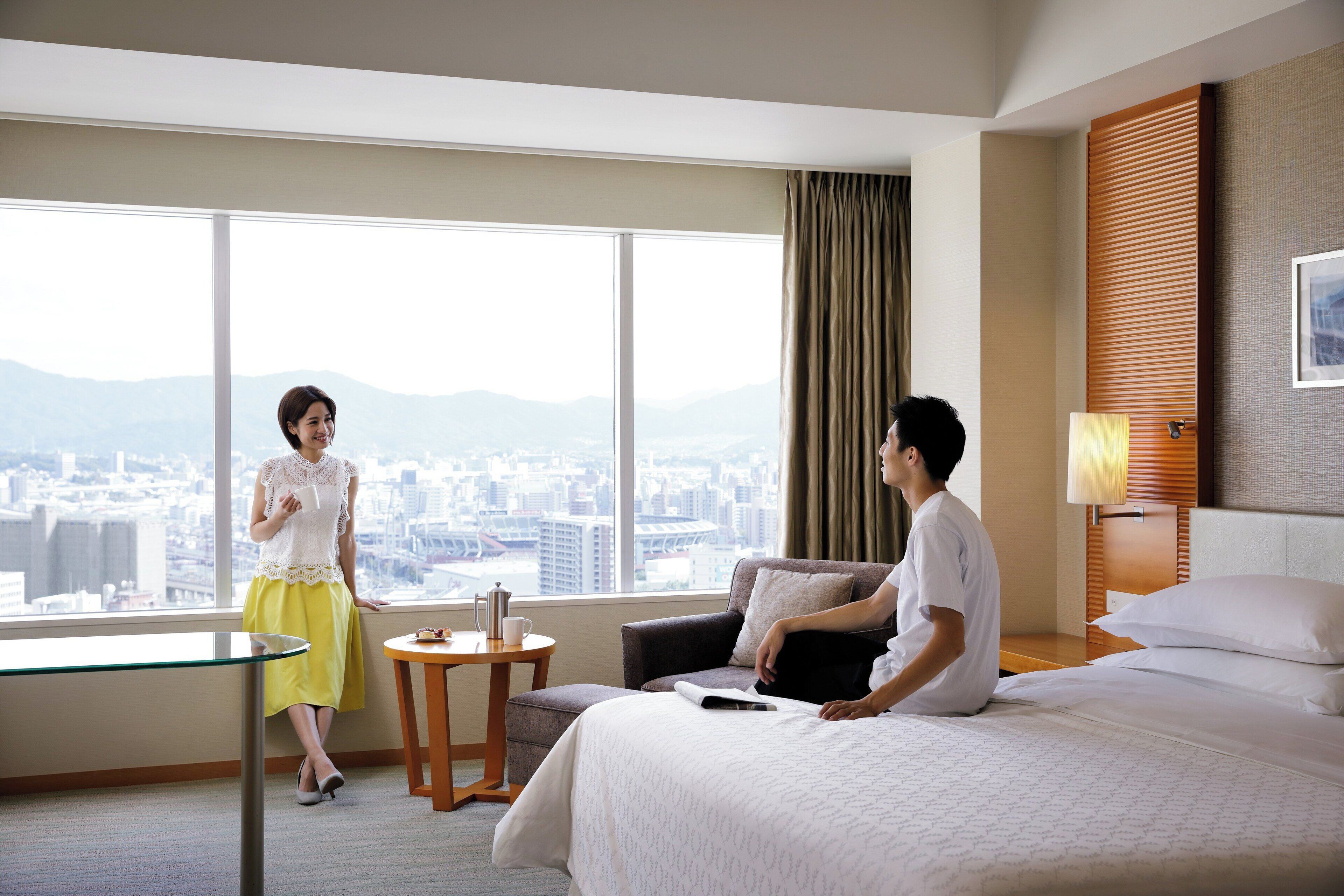 A hotel room with a large window, a bed, a small table, and a sofa.