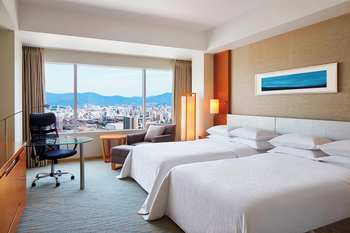 A hotel room with two beds, a desk, a chair, a lamp, and a large window offering a city view.