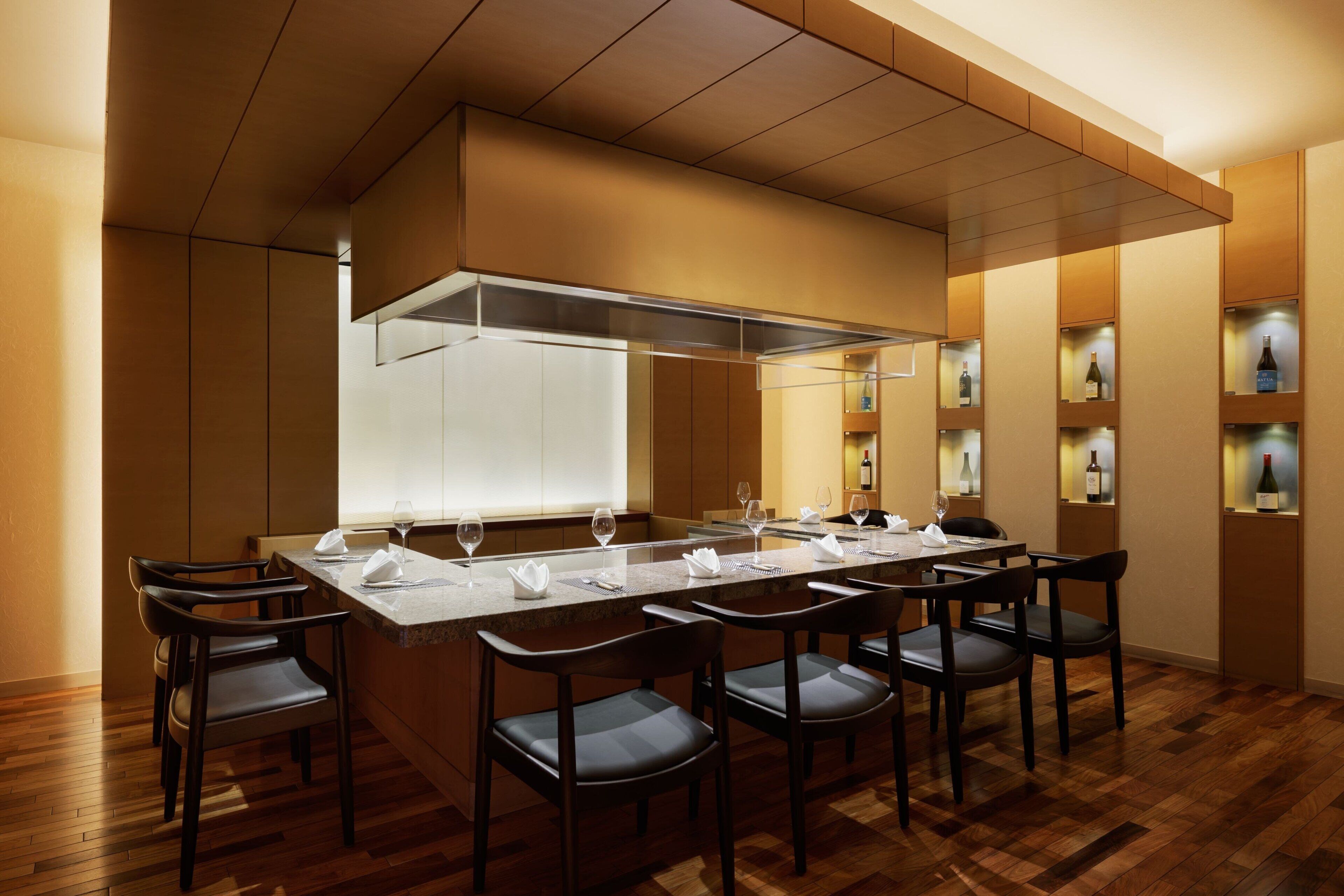 A restaurant with a long table set for six, wooden flooring, and a modern kitchen visible in the background.