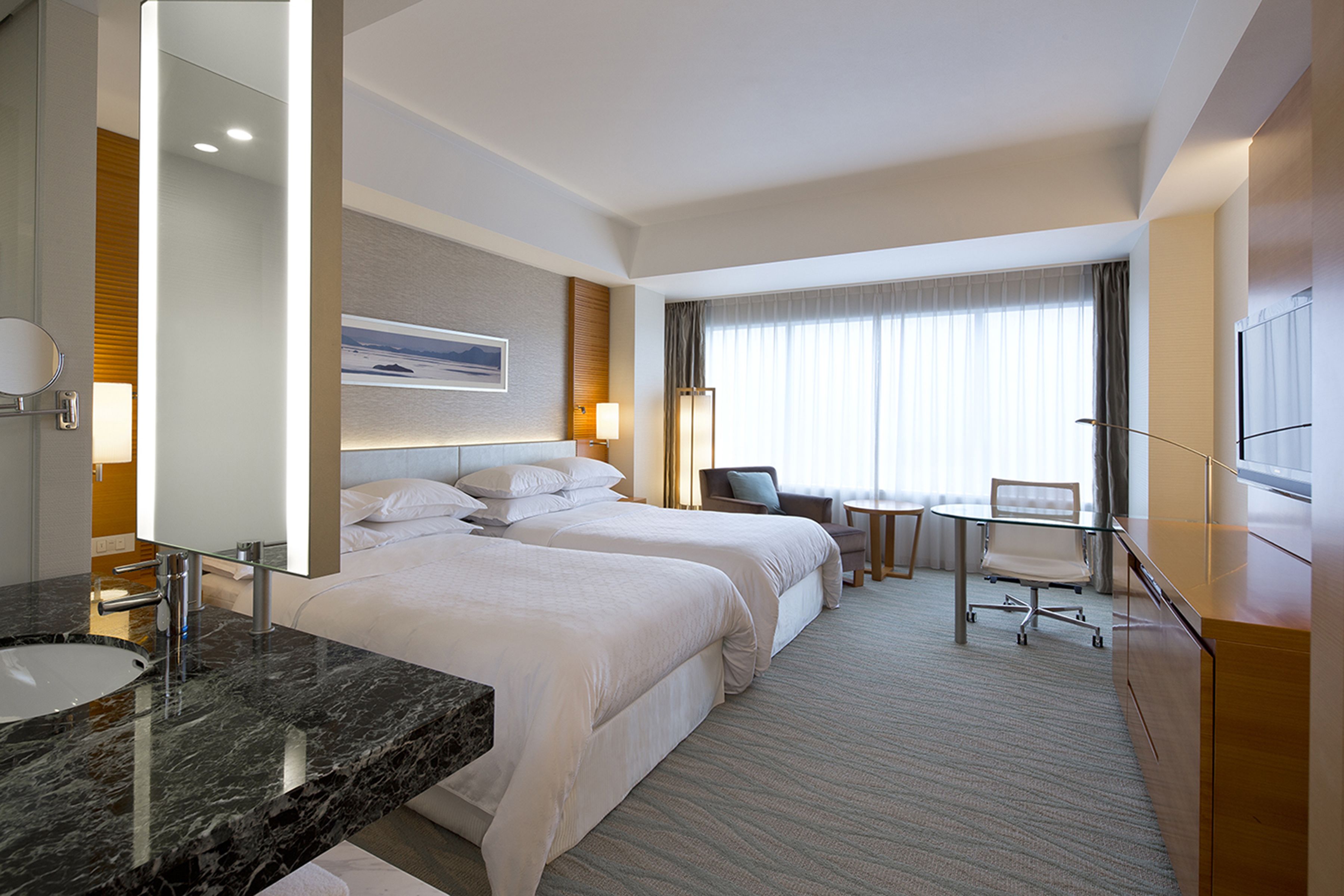 Habitación de hotel con lavamanos de mármol, dos camas, un escritorio y un ventanal con cortinas.