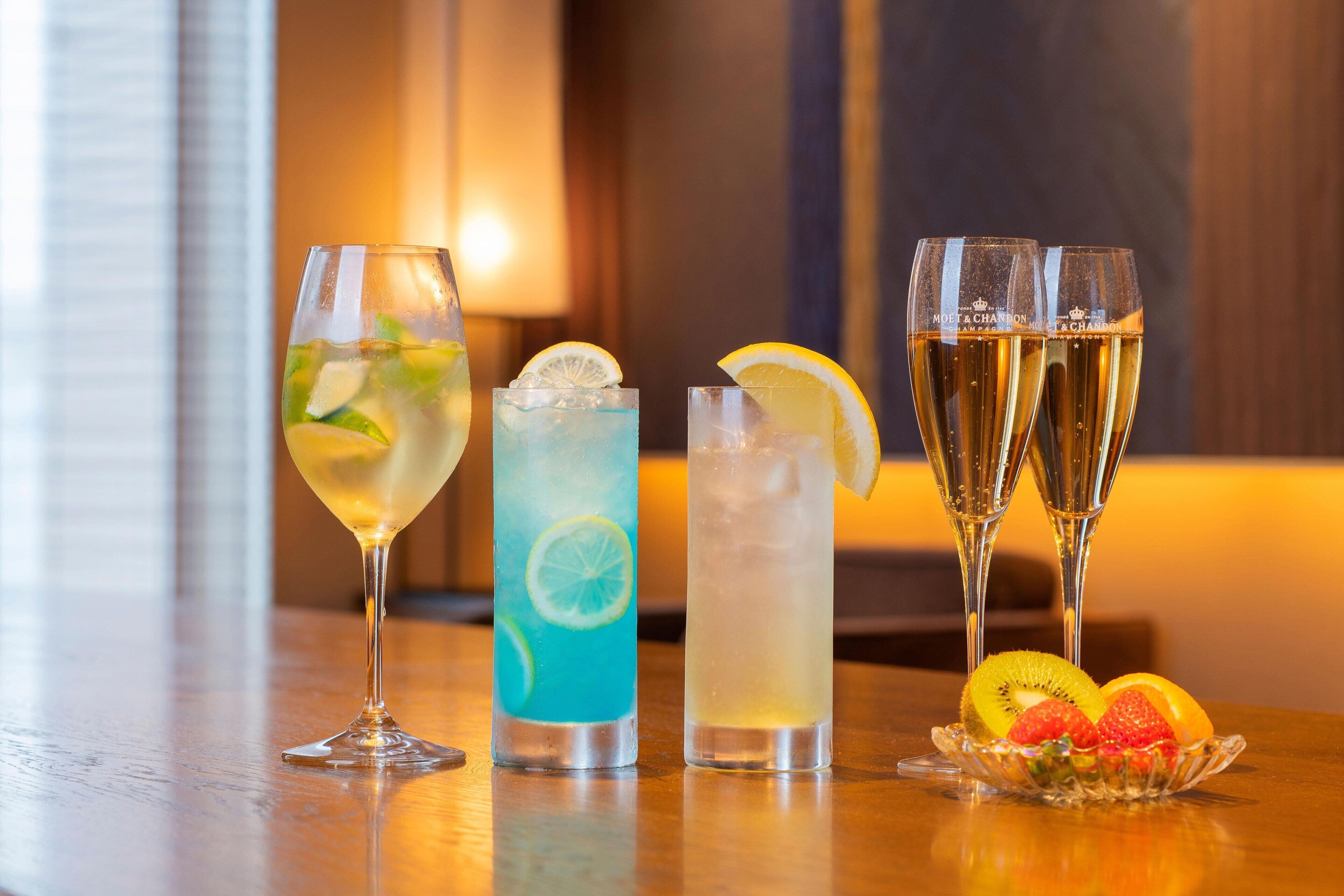 Three glasses of beverages on a wooden table with a fruit bowl containing strawberries and kiwi.