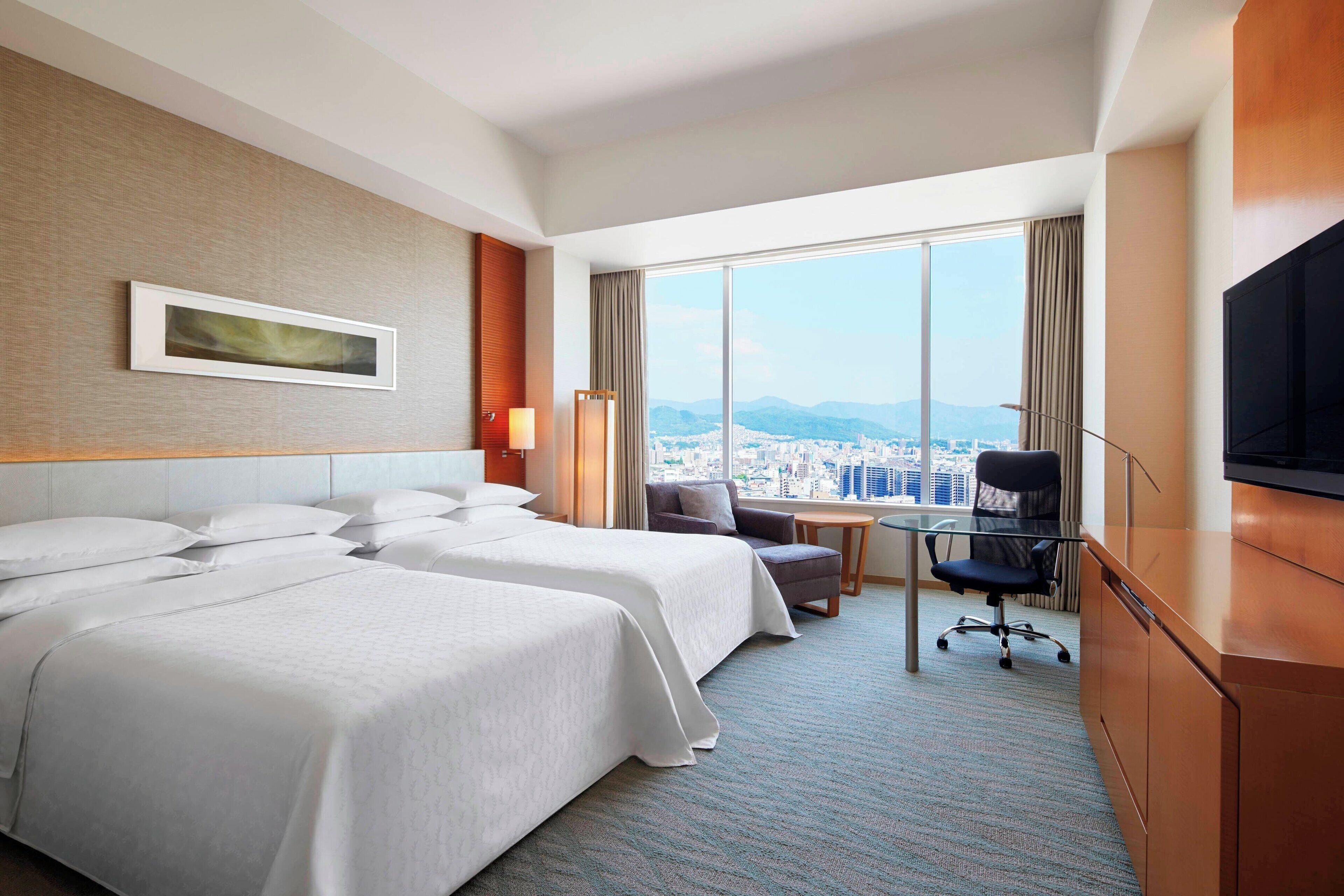 A hotel room with two beds, a desk, a chair, a television, and a large window offering a city view.