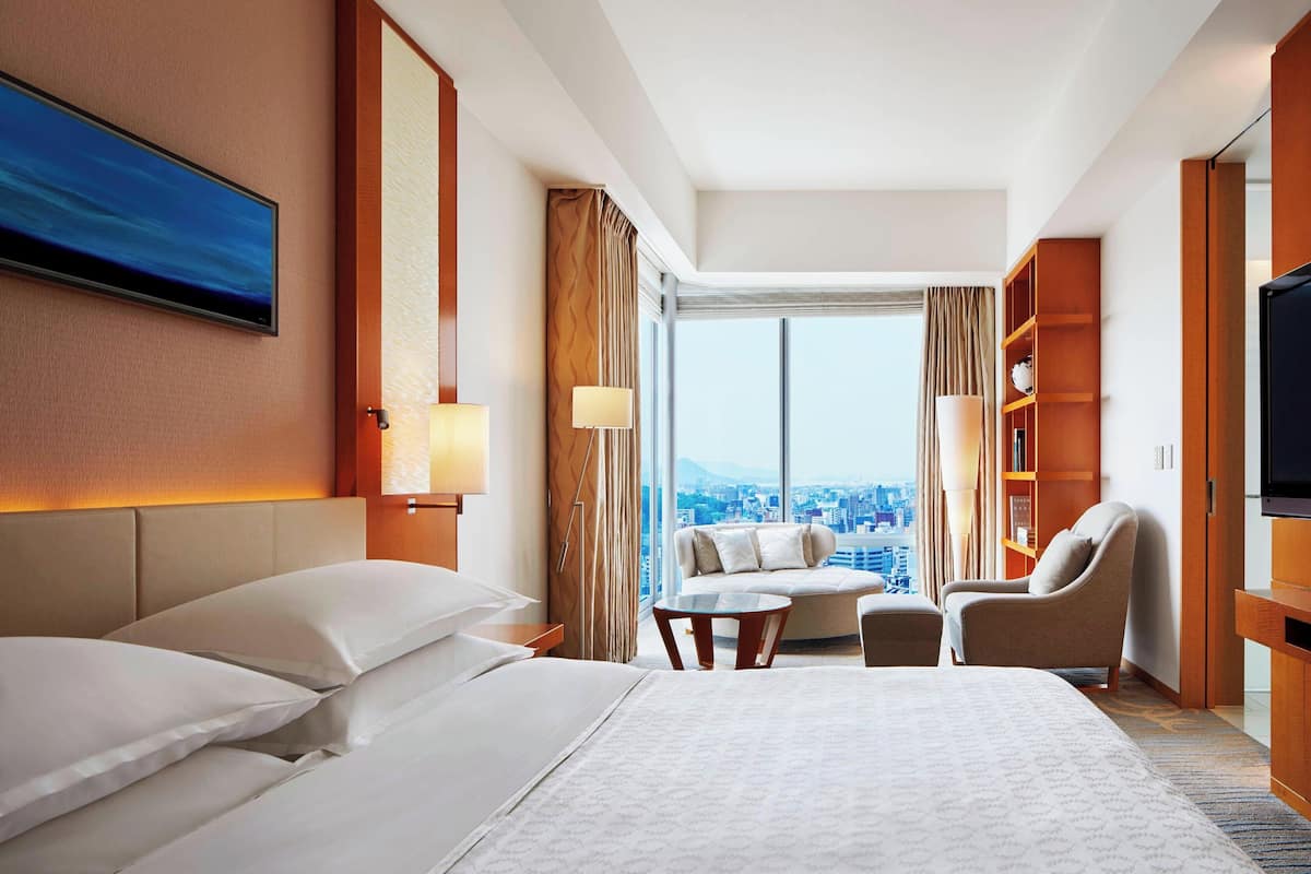 A hotel room with a large bed, a sofa, a TV, and a view of a cityscape.